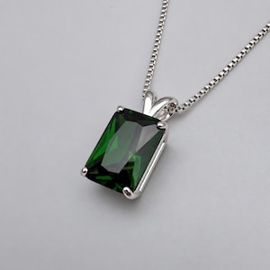 May include: A silver necklace featuring a rectangular, emerald-cut dark green gemstone pendant held by a four-prong setting. The pendant is attached to a delicate silver box chain that extends upwards from the top of the metal mounting.