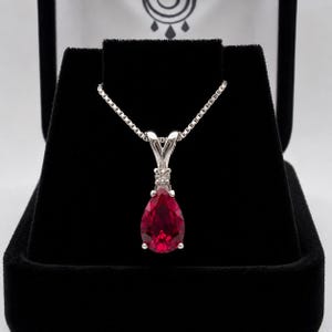 May include: A silver necklace featuring a pear-shaped, deep red gemstone pendant suspended from a delicate box chain. The pendant is accented with a small, clear round stone set in silver above the main red teardrop-shaped gemstone.