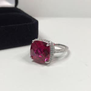 May include: A silver ring with a large, square-shaped, red gemstone surrounded by a halo of smaller, white gemstones.