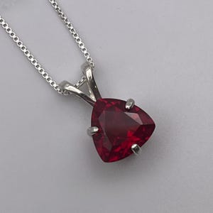 May include: A silver necklace with a triangular, faceted, deep red pendant. The pendant is held in place by four silver prongs. The chain is a delicate, linked design. The background is a light gray.