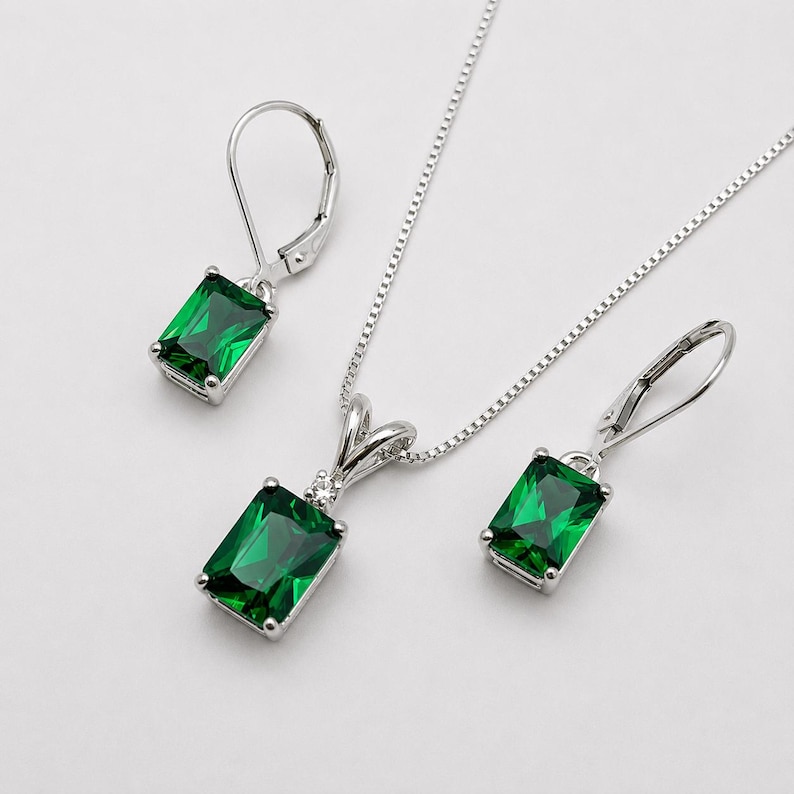 May include: A jewelry set consisting of a silver necklace and two matching leverback earrings. Each piece features a rectangular emerald green gemstone set in silver, with the pendant accented by a small clear stone at the top bail.