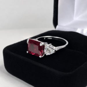May include: A silver ring featuring a large, square-cut, deep red gemstone flanked by two smaller, triangular-cut, clear white stones. The inner band is clearly stamped with the hallmark .925, indicating sterling silver construction.