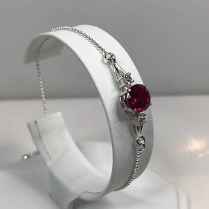 Pode incluir: Uma pulseira de prata com uma pedra preciosa vermelha e duas pedras preciosas transparentes menores. A pulseira tem uma corrente delicada.