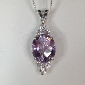 May include: Silver pendant necklace featuring a large, oval-cut, light purple gemstone. The gemstone is surrounded by smaller, clear, round stones. The pendant hangs from a delicate silver chain. The jewelry is set against a white background.