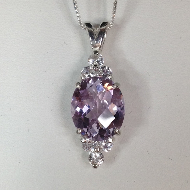 May include: Silver pendant necklace featuring a large, oval-cut, light purple gemstone. The gemstone is surrounded by smaller, clear, round stones. The pendant hangs from a delicate silver chain. The jewelry is set against a white background.