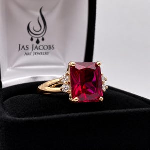May include: A gold ring featuring a large rectangular red gemstone flanked by three small round clear stones on each side. The ring is presented in a black jewelry box with the text JAS JACOBS ART JEWELRY printed on the white interior lining.