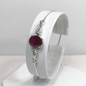 May include: A silver bracelet with a red oval gemstone and smaller clear gemstones. The bracelet has a sliding clasp.