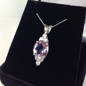 May include: A silver necklace with a large, oval, light purple gemstone pendant. The pendant is surrounded by smaller, clear gemstones. The necklace is displayed on a black velvet surface.