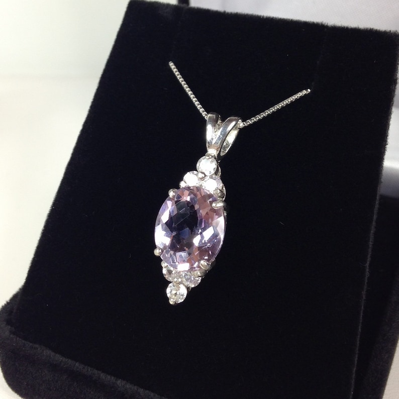 May include: A silver necklace with a large, oval, light purple gemstone pendant. The pendant is surrounded by smaller, clear gemstones. The necklace is displayed on a black velvet surface.