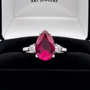 May include: A silver-toned ring featuring a large, pear-shaped, deep pink gemstone as the centerpiece, flanked by two smaller, round, clear stones on either side. The ring is set against a black velvet interior of a jewelry box.