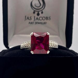 May include: A silver ring with a large, rectangular, deep red gemstone. The band features a detailed leaf design. The ring is displayed in a black jewelry box with the text "Jas Jacobs Art Jewelry".