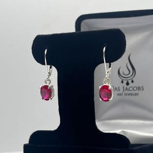 May include: A pair of silver dangle earrings with red oval-shaped gemstones. The earrings are displayed on a black velvet background with a white box that says "Jas Jacobs Art Jewelry".