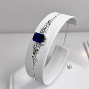 May include: A silver chain bracelet featuring a central rectangular deep blue gemstone flanked by two small clear round stones. The bracelet has a delicate box chain design and a polished metallic finish, showcasing a refined and symmetrical arrangement.