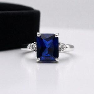 May include: A silver-toned ring featuring a large, rectangular, deep blue center stone held by four prongs. The band is accented on each side by a single, small, round, clear stone set into the metal, creating a three-stone design.