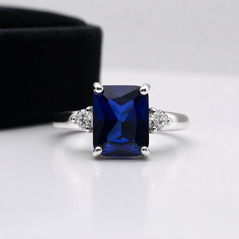 May include: A silver-toned ring featuring a large, rectangular, deep blue center stone held by four prongs. The band is accented on each side by a single, small, round, clear stone set into the metal, creating a three-stone design.