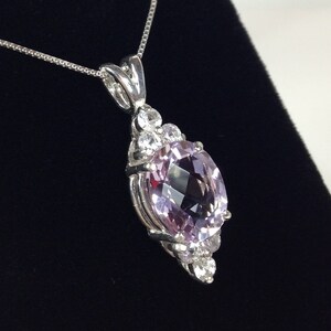 May include: A silver pendant necklace featuring a large, oval-cut, light purple gemstone. The gemstone is surrounded by smaller, clear gemstones. The pendant hangs from a delicate silver chain, set against a black background.