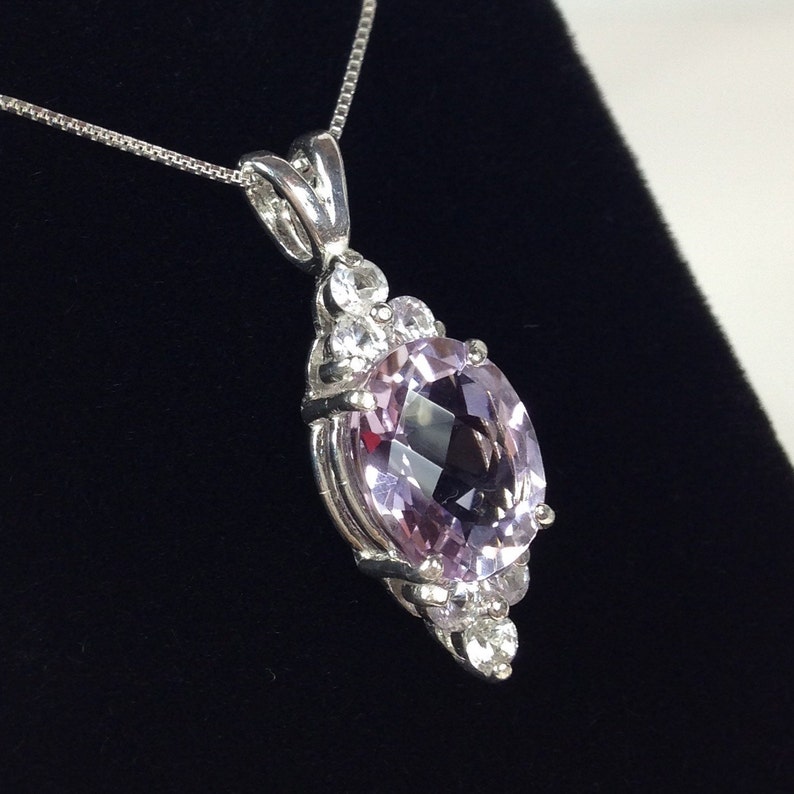 May include: A silver pendant necklace featuring a large, oval-cut, light purple gemstone. The gemstone is surrounded by smaller, clear gemstones. The pendant hangs from a delicate silver chain, set against a black background.