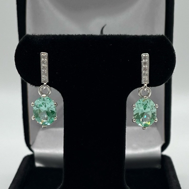 May include: A pair of silver dangle earrings with a light green gemstone and a row of clear stones on the top of the earring.