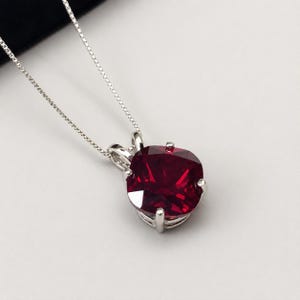 May include: A silver necklace featuring a cushion-cut deep red gemstone pendant held by four prongs. The pendant is suspended from a delicate silver box chain. The gemstone has a faceted surface that reflects light, creating a bright appearance.
