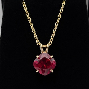 May include: A gold chain necklace featuring a cushion-cut, deep red gemstone pendant. The vibrant red stone is secured by four gold prongs in a classic setting, suspended from a delicate gold link chain that hangs vertically against a dark background.