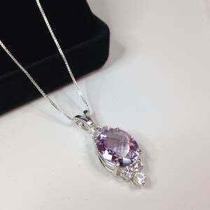May include: A silver necklace featuring a pendant with a large, oval-cut, light purple gemstone. The pendant is accented with smaller, clear gemstones. The necklace is displayed on a white surface with a black velvet box in the background.
