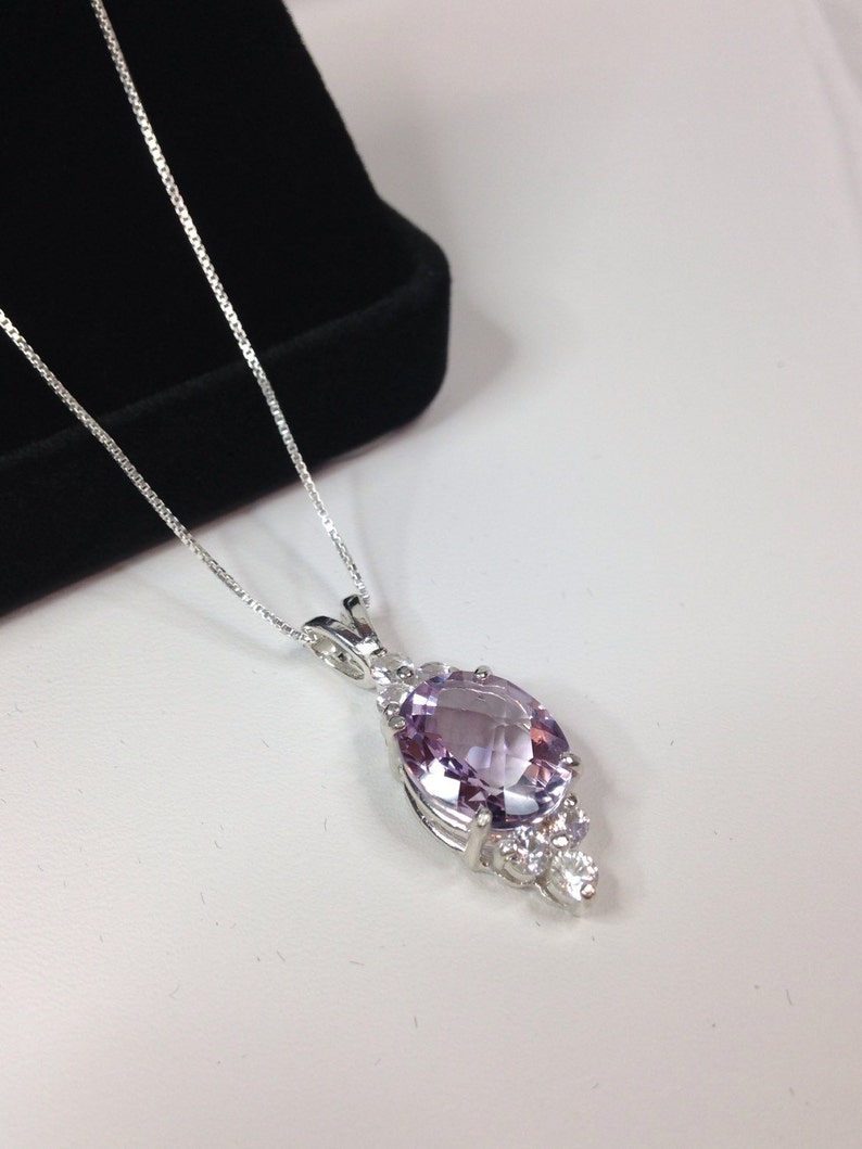 May include: A silver necklace featuring a pendant with a large, oval-cut, light purple gemstone. The pendant is accented with smaller, clear gemstones. The necklace is displayed on a white surface with a black velvet box in the background.