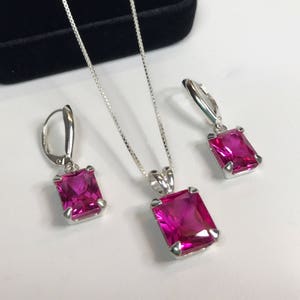 May include: A jewelry set featuring a silver necklace with a rectangular, magenta-colored pendant, and matching leverback earrings. The earrings and pendant have a silver setting. The set is displayed on a white surface.
