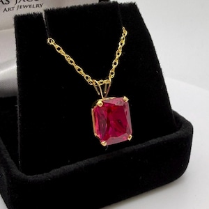 Puede incluir: Un collar de oro con un gran colgante rectangular de piedra preciosa rojo rubí. El colgante está engastado en un marco de oro y cuelga de una delicada cadena de oro. El collar se exhibe en una caja de joyería negra.