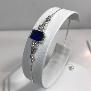 May include: A silver bracelet with a blue gemstone and clear accents. The bracelet has an adjustable sliding clasp.
