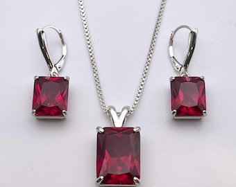 May include: A silver-toned jewelry set featuring a pendant necklace and matching earrings. Each piece showcases a rectangular, deep red gemstone set in a silver frame. The earrings have lever-back closures.