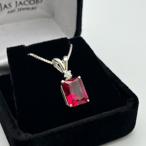 Puede incluir: Un collar de plata con un colgante de piedra roja y un pequeño acento de piedra clara. El collar está en una caja de terciopelo negro con las palabras "Jas Jacobs Art Jewelry" en la tapa.