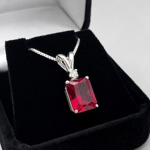 May include: A silver pendant necklace featuring a large, rectangular, deep red gemstone. The stone is set in a silver frame with a smaller, round, clear accent stone positioned above it, suspended from a delicate silver box chain.