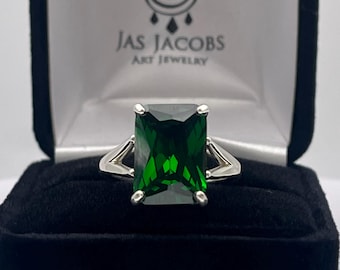 May include: A silver ring with a large emerald green gemstone in an emerald cut setting. The ring is displayed in a black velvet ring box with the words "Jas Jacobs Art Jewelry" printed on the inside of the lid.
