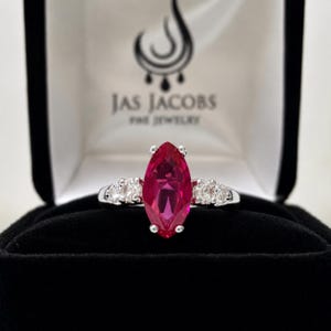May include: A silver ring featuring a large marquise-cut vibrant red gemstone at the center, flanked by two small round clear stones on each side. The ring is presented in a black jewelry box with the text Jas Jacobs Fine Jewelry printed inside.