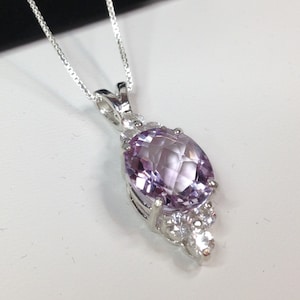 May include: A silver necklace with a pendant featuring a large, oval-cut, light purple gemstone. The pendant is accented with smaller, clear gemstones and hangs from a delicate silver chain. The pendant is approximately 1 inch in length.