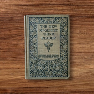 May include: The cover of a vintage book titled "The New McGuffey Third Reader" with an ornate floral design in black and gold. The book was published by the American Book Company in New York, Cincinnati, and Chicago.