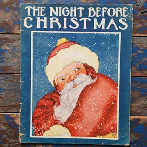 May include: A vintage book cover featuring a blue background with white snowflakes. The cover has the title "The Night Before Christmas" in bold, white letters. The illustration shows Santa Claus in a red suit and white beard, looking over his shoulder with a mischievous grin.