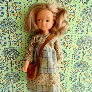 May include: A doll wearing a brown and blue plaid dress with a brown leather strap purse. The doll has blonde hair and blue eyes. The doll is standing in front of a green and blue floral patterned background.