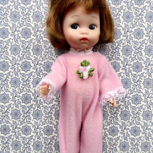 May include: A vintage doll with brown hair and large eyes, wearing a pink onesie with white lace trim and a floral applique. The doll is posed against a blue and white floral patterned background.