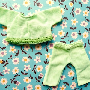 May include: A light green doll-sized outfit, including a short-sleeved top and pants. The top has a decorative trim along the bottom. The outfit is made of a ribbed fabric. The background is a blue floral pattern.