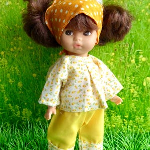 May include: A doll with brown hair styled in two pigtails, wearing an orange headscarf with white polka dots, a white shirt featuring a small yellow carrot pattern, and solid yellow pants with matching carrot-patterned cuffs and yellow shoes.