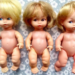 May include: Three vintage baby dolls with blonde hair and blue or brown eyes. The dolls are nude, with a pinkish skin tone. They are posed on a light blue floral patterned surface.