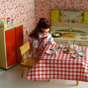 May include: A dollhouse kitchen set with a red and white checkered tablecloth, a doll sitting at the table, and a dollhouse stove with a clock and a red and white checkered countertop.