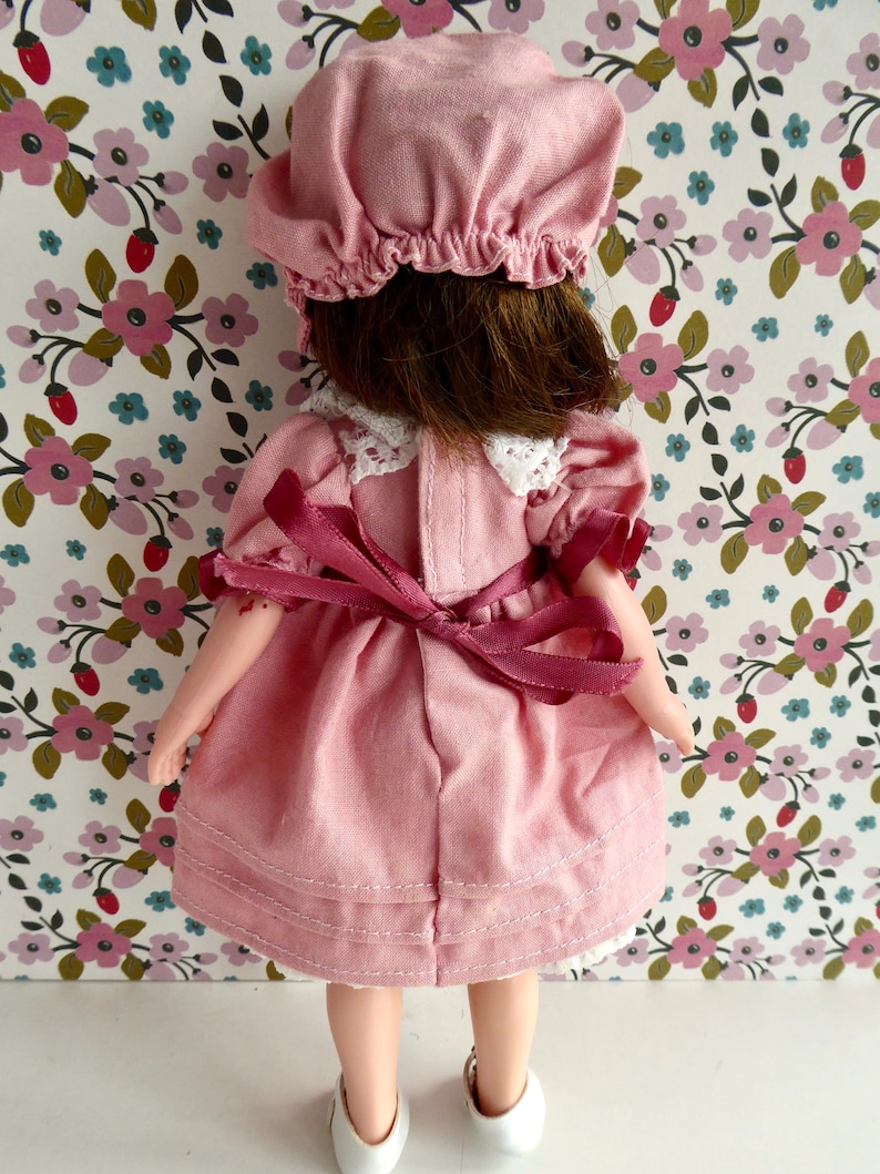 Op de afbeelding: Een pop in een roze jurk met korte pofmouwen, een witte kanten kraag en een donkerroze lint dat op de rug is gestrikt. De pop heeft bruin haar en draagt een bijpassend roze mutsje. De jurk heeft een geplooide rok en witte schoenen.