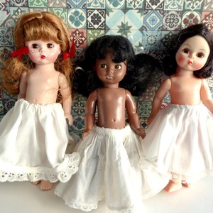 May include: Three dolls wearing white petticoats with lace trim. The dolls have different skin tones and hair colours. The dolls are posed against a background of colourful tiles.