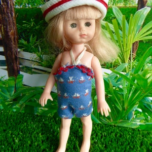 May include: A doll with blonde hair wearing a blue and white swimsuit with a red trim and a white and red sailor hat. The doll is standing on a green grassy surface.