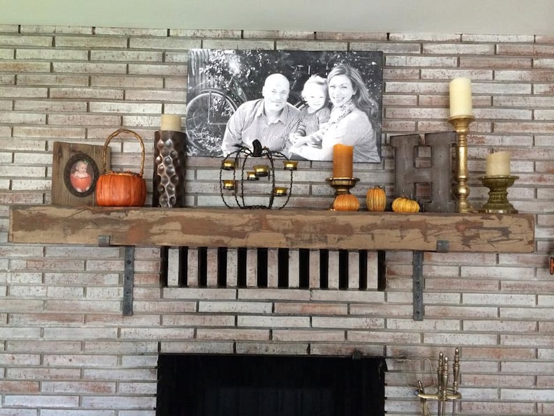 May include: A rustic wooden mantel with a black and white photo of a family, a metal pumpkin candle holder, and small pumpkins. The mantel is decorated with candles, a metal letter H, and a small picture frame.