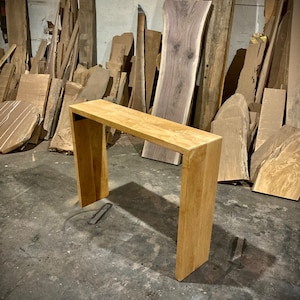 May include: A light brown wooden console table with a rectangular top and two vertical supports. The table is standing on a concrete floor in a workshop setting.