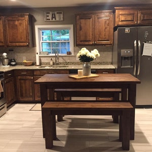 May include: A wooden table and bench set in a kitchen with brown cabinets and a stainless steel refrigerator. The table and bench are both made of dark wood and have a rustic look. The table has a simple design with a rectangular top and four legs. The bench has a similar design with a long seat and two legs on each end. The table and bench are both in good condition and would be a great addition to any home.