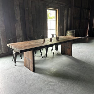 May include: A rustic wooden table with a natural wood finish sits in a room with exposed wooden beams and a large window. Four black metal chairs surround the table.